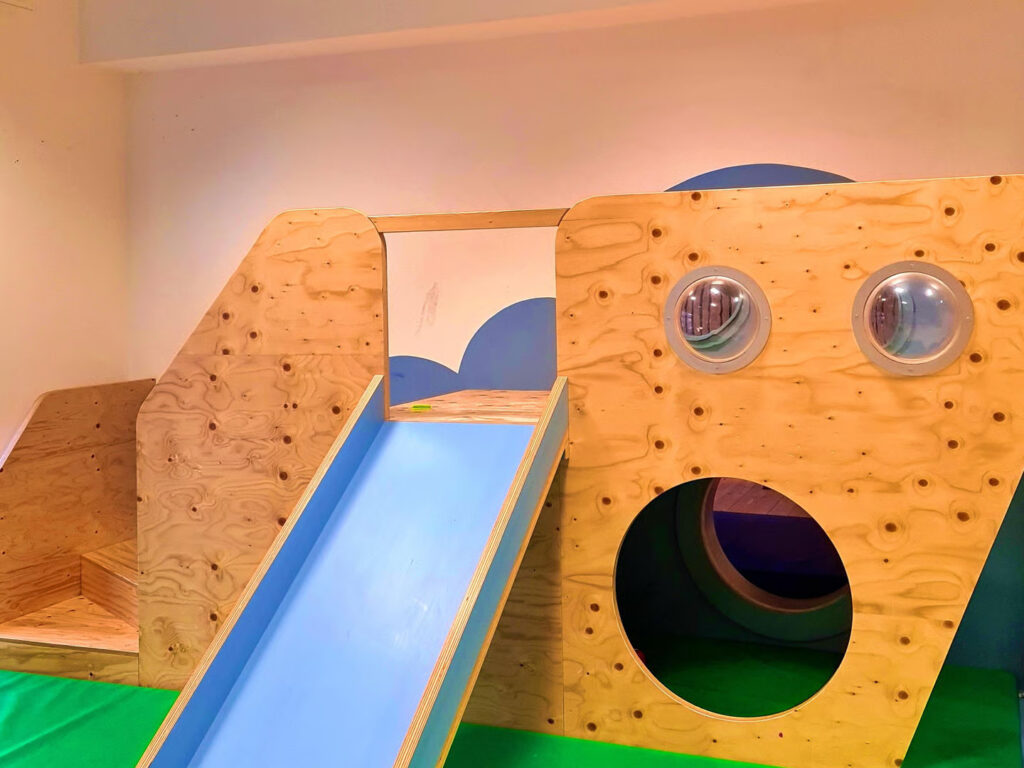 Ein Holzspielhaus mit blauer Rutsche Bullaugen und rundem Krabbeltunnel